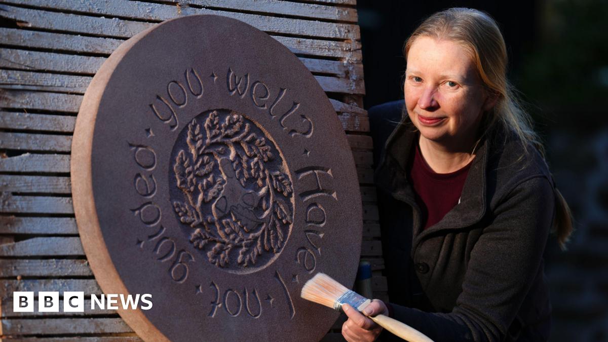 Jennie is on the right of the picture, looking at the camera, while leaning around a pallet-like piece of wood. She's smiling at the camera, and holding a paintbrush, wearing a black jumper. Next to her is a stone circle with an inscription and intricate design.