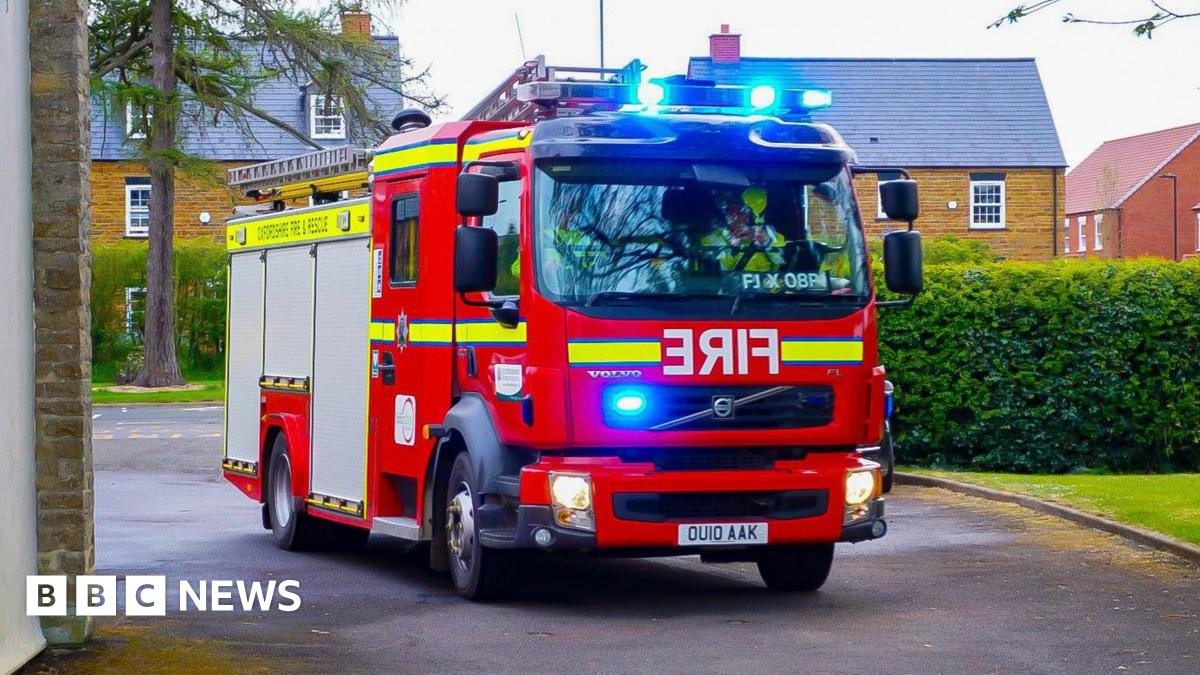 A new-looking fire engine, bright red in colour, faces the camera at a slight angle. Emergency lights glow blue at the top and front of the vehicle. Behind the engine you can see greenery and some residential homes.