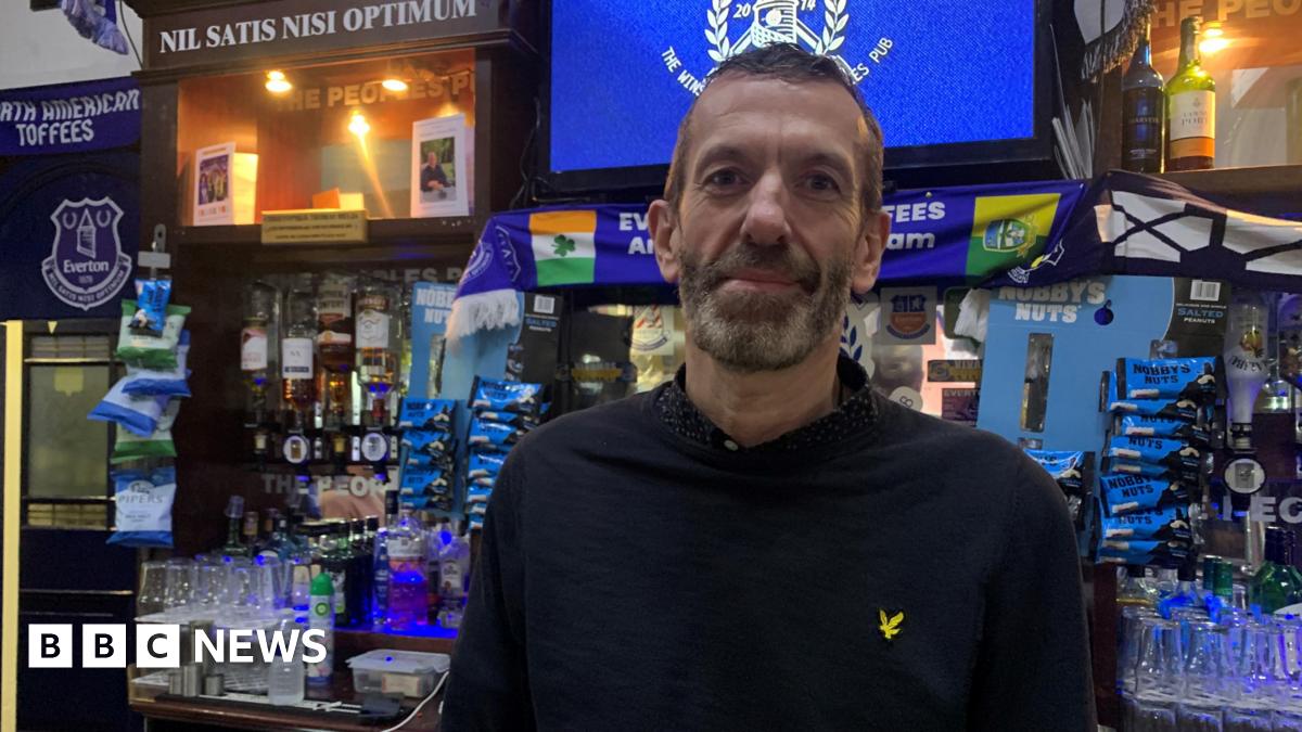 Dave stands in front of an adorned bar filled with Everton scarves. He is wearing a dark polo shirt and has short dark hair