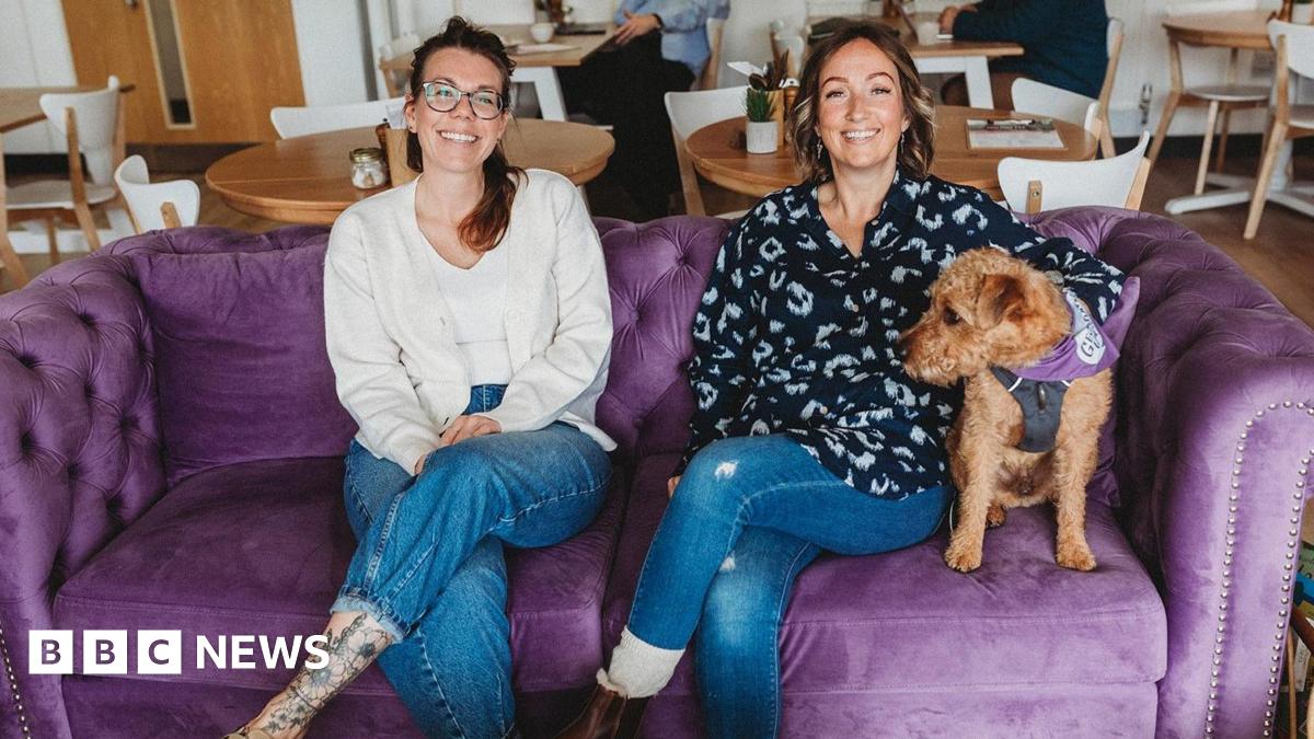 Kirsty, a trustee, and Sophie are sitting on a plush velvet couch in the cafe. Sophie (on the right) has a small, brown curly-haired dog sitting under her arm. Both are smiling broadly and have their legs crossed. Two people can be seen sitting at tables behind them.