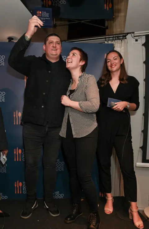 Getty Images Chef Gareth Ward stands on a small stage in front of a “The Good Food Guide” backdrop. He raises an award plaque above his head while smiling, his guest, a woman, looks up at it, smiling, and a woman on the right smiles while holding a microphone and a card.