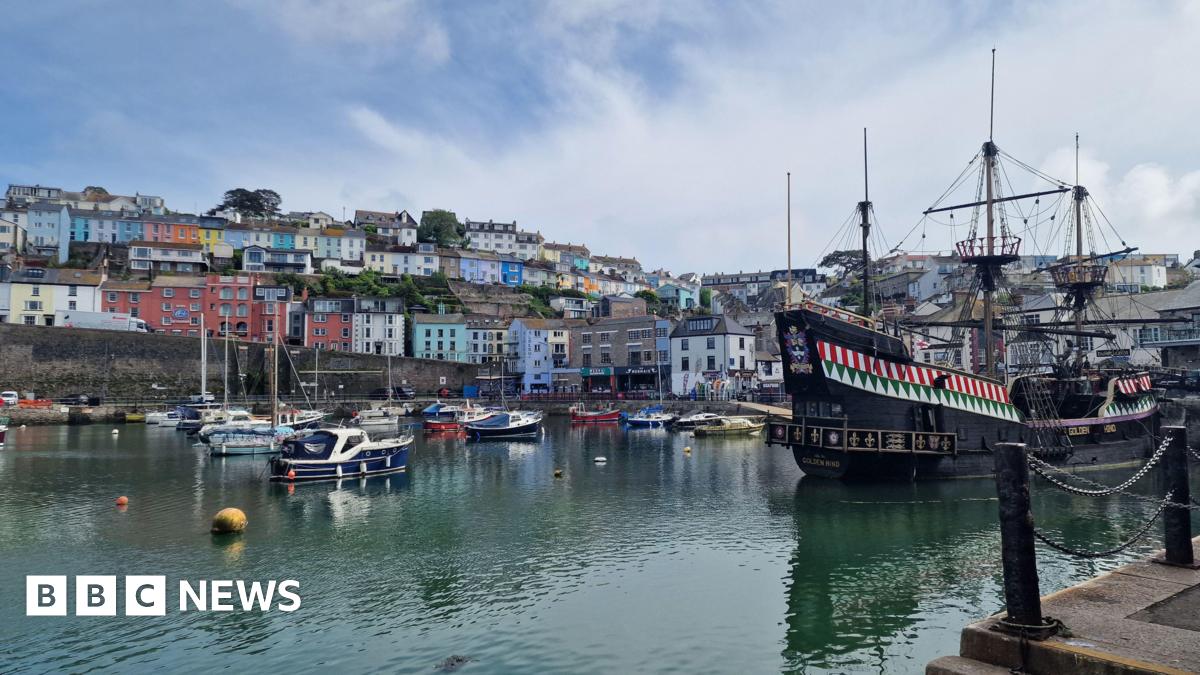 Boats in a harbour with calm waters. Colourful properties built near the harbour overlook the water. A large wooden ship is also moored in the harbour.