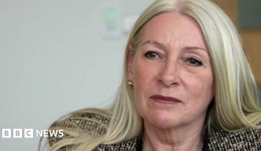 Kay Mason Billig, who has blonde hair, is facing the camera but looking to the left of the frame. She's wearing a patterned suit jacket.