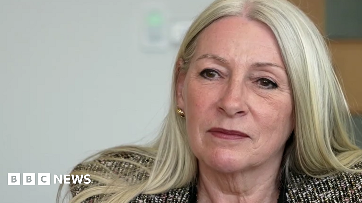 Kay Mason Billig, who has blonde hair, is facing the camera but looking to the left of the frame. She's wearing a patterned suit jacket.