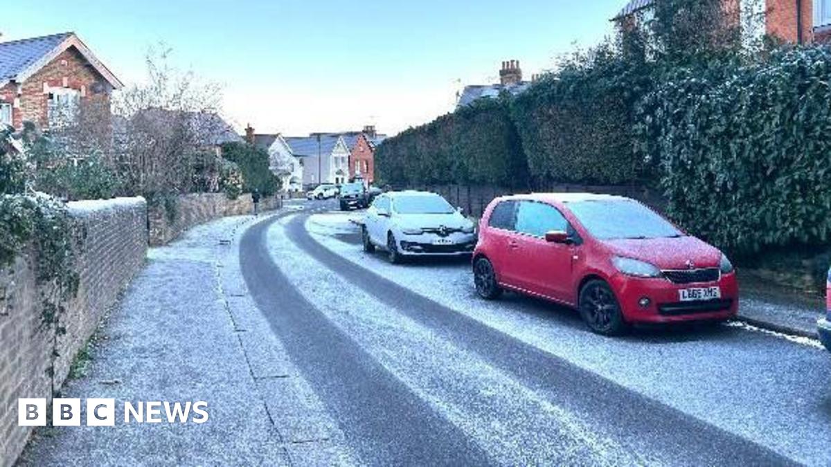 A small residential street with light dustings of snow on it. A red vehicle is parked in front of a white vehicles, facing towards the camera, on the opposite side of the road