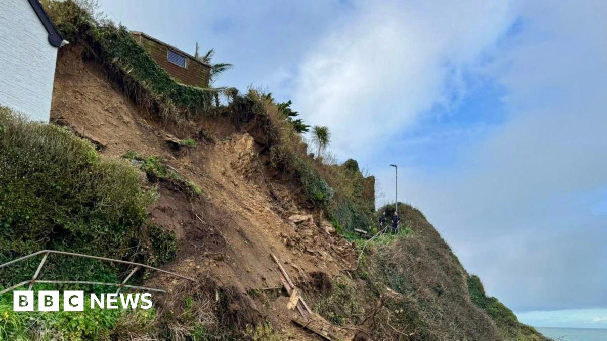 Part of a cliff seen collapsed on the side of the hill, a small building sits at the top and two people are seen in the distance on a path.