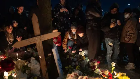Getty Images People gather at a memorial for Good. there are candles, flowers and a big cross.