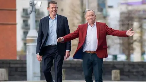 Getty Images Rhun ap Iorwerth with successful Plaid Cymru candidate Lindsay Whittle, stood in Cardiff Bay with a slate wall behind them. Whittle is wearing a red jacket. blue trousers and white shirt and has his hands open. Ap Iorwerth is smiling towards him.