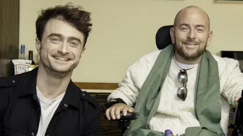 Issac Lamb Daniel Radcliffe wearing a white top and black jacket next to David who is wearing a cream jumper and is in a wheelchair. They are both smiling, sitting inside.