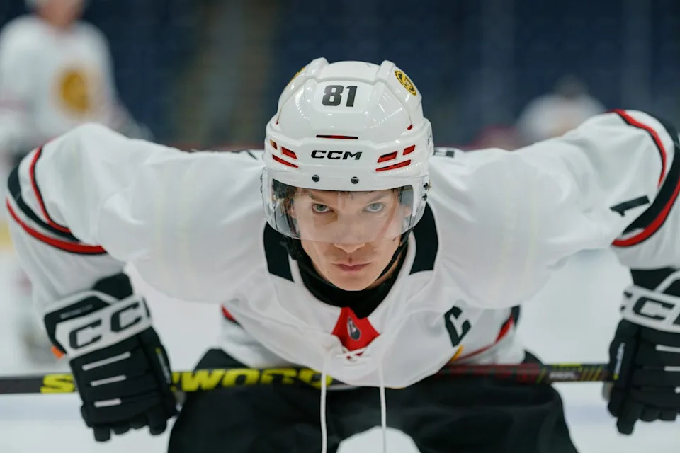 A white man in a hockey uniform leans over while holding his stick against his thighs