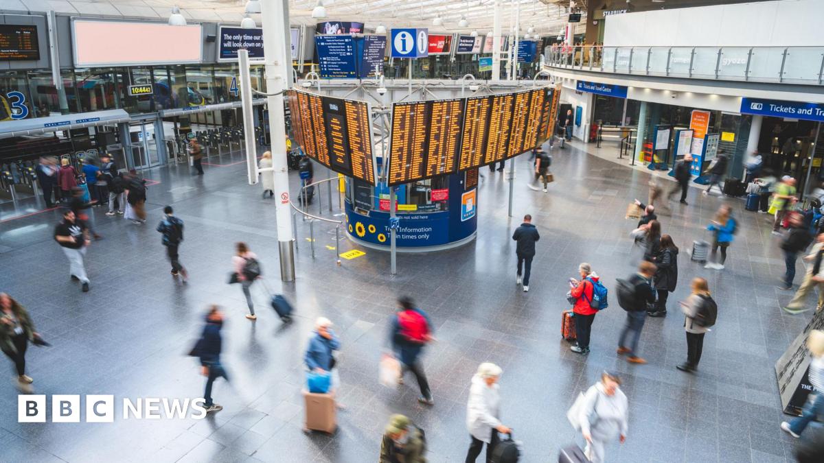 An overhead view of Manchester Piccadilly station and a central departures board lit up with train destinations and times. Around thirty people are crossing the concourse, blurred in the photo due to movement.