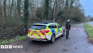 A police officer stands next to a patrol car, with a taped off area behind him.