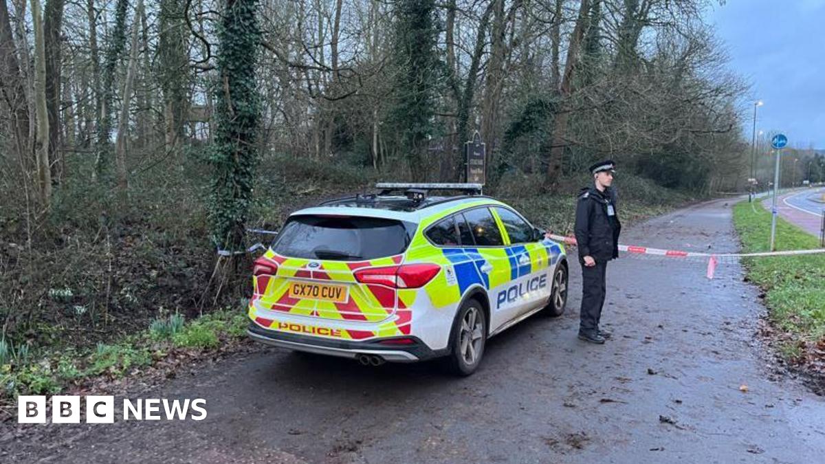 A police officer stands next to a patrol car, with a taped off area behind him.