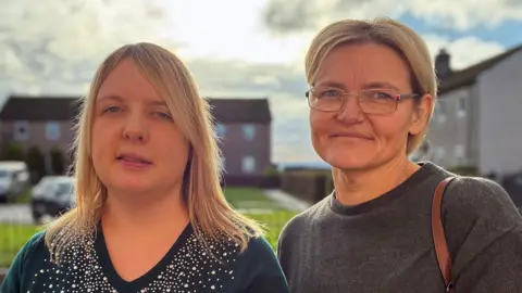 BBC Two women with blonde hair, one on the right wearing glasses, looking directly at the camera with houses in the background