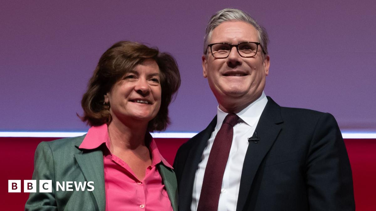 A woman with brown hair, green suit jacket and pink shirt smiles while standing next to a man with grey hair and glasses, wearing a navy suit, white shirt and pink tie.