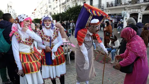APP/NurPhoto via Getty Images Two women, wearing face paint and colourful wigs, stand by a masked figure.