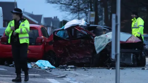 Phil Taylor Crash scene shows two badly damaged cars and debris in the road. Two police officers wearing yellow hi-vis jackets are at the scene.