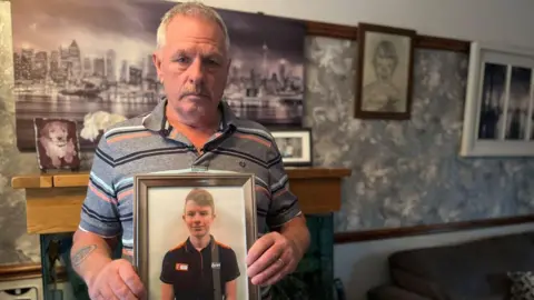 BBC A man with grey hair and a grey polo top holds up a picture of a young man. The photo of the older man is taken in his living room