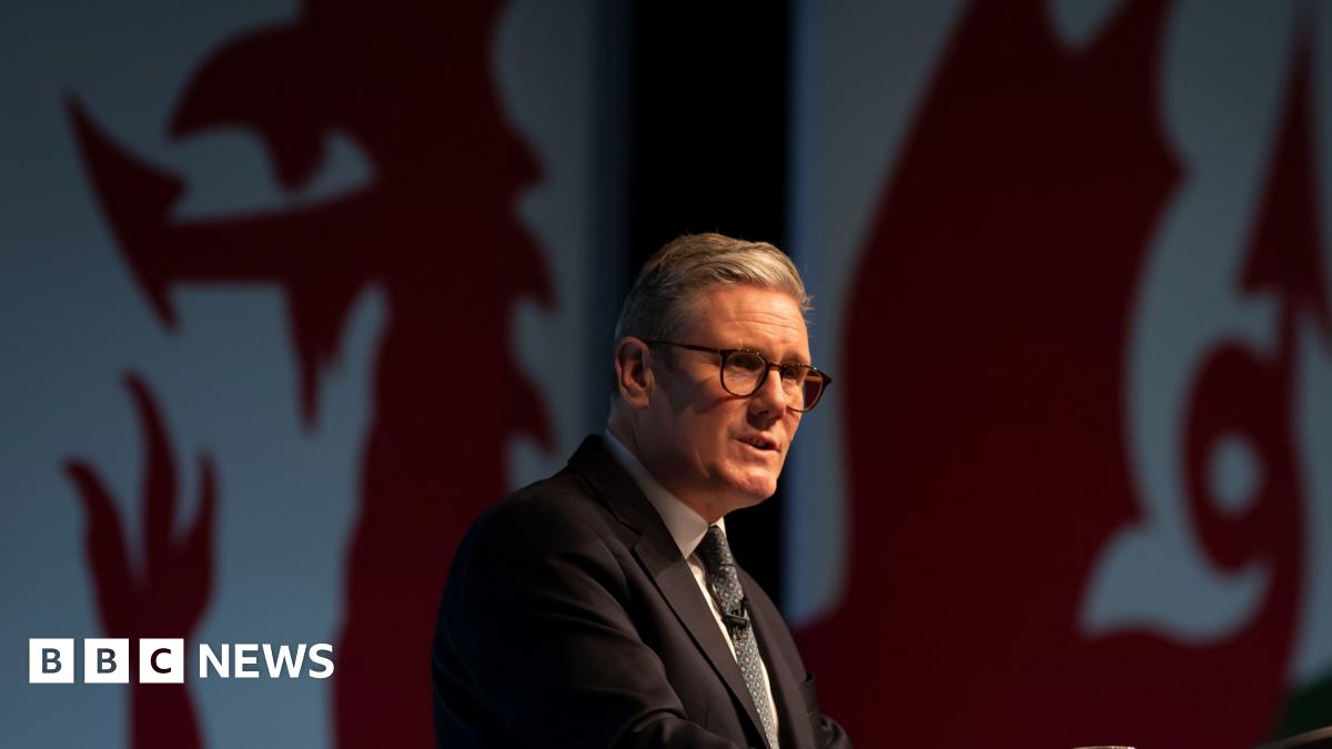 A man with grey hair, glasses, a navy suit, blue shirt and a red tie - Sir Keir Starmer - stands at a podium while giving a speech. In the background sits a woman with medium length brown hair wearing a white suit - Eluned Morgan - alongside two women and a man.