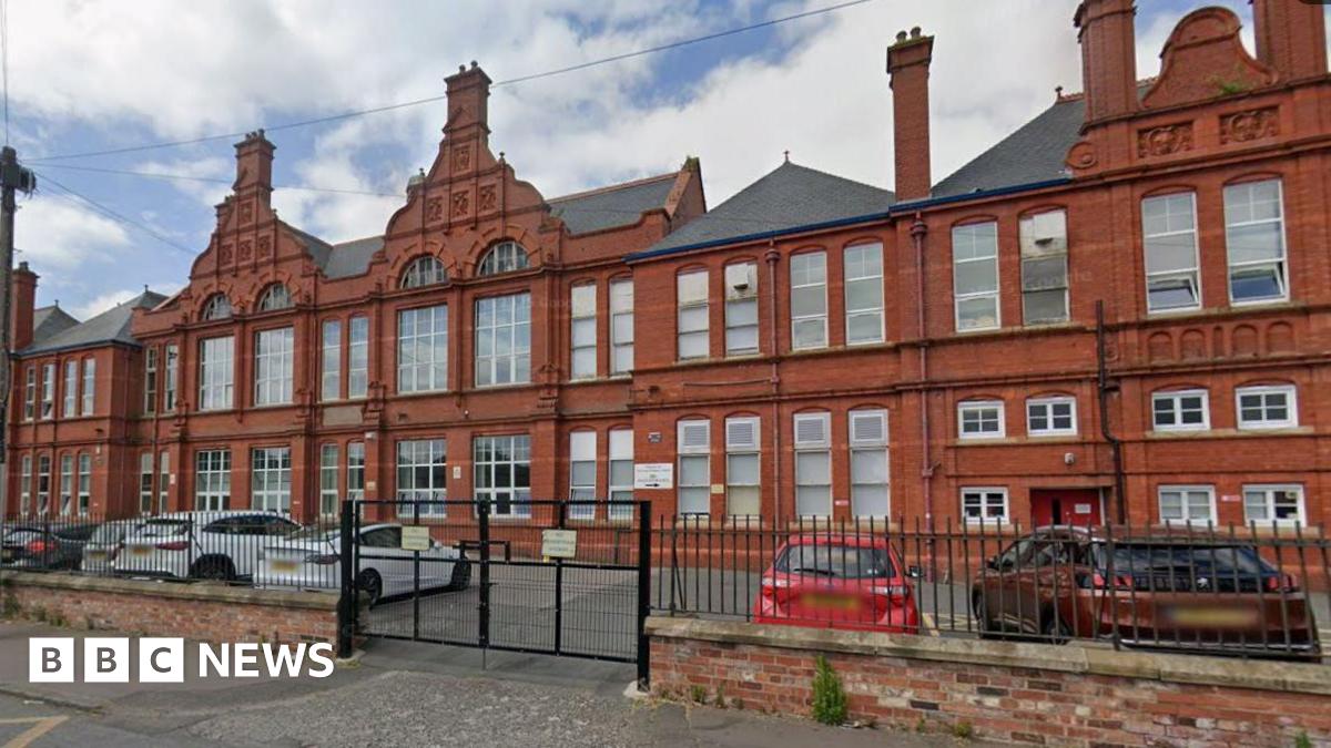 A red brick school building of three stories, with a car park in front and several cars parked there. There are railings around it which are closed.