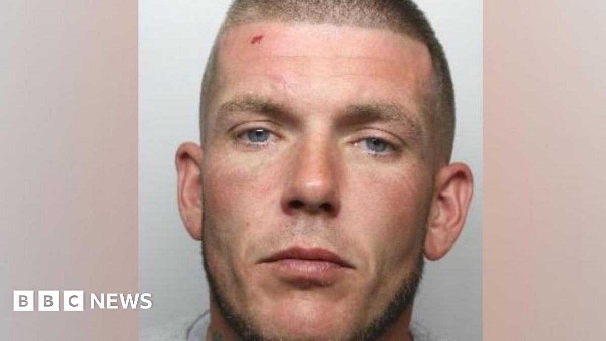 A custody photo shows a man with a shaved head and blue eyes and a cut on his forehead. He is wearing a grey t-shirt.