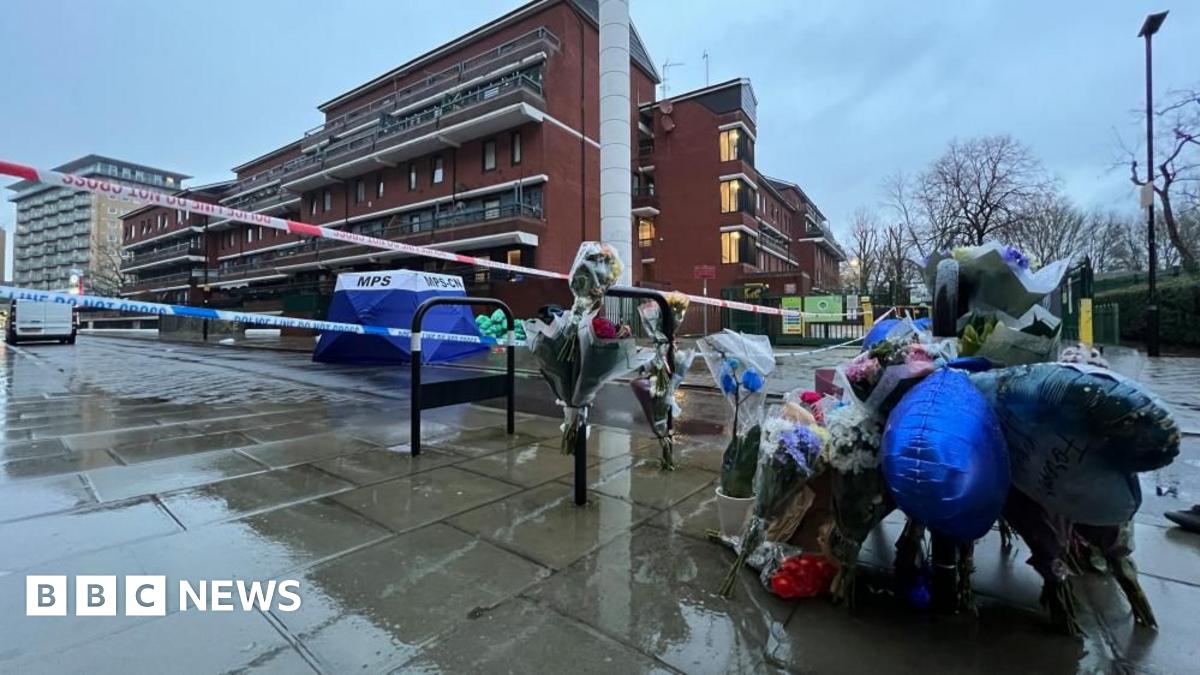 A number of flowers are left at a rainy looking crime scene with a blue MPS tent stationed in the background. Police tape lines the area.