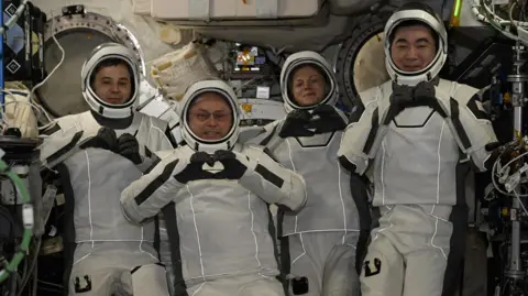 NASA Four astronauts wearing white spacesuits and helmets pose for cameras smiling and making heart shapes with their hands and fingers. They are surrounded by wires and other equipment inside the International Space Station.