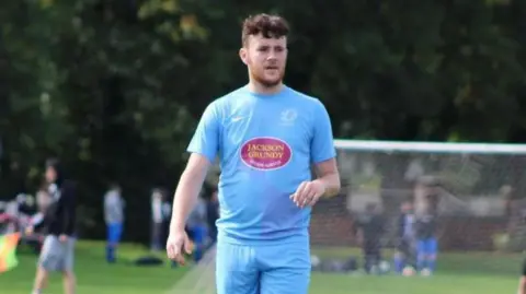 Richard Abrahams  Luke Abrahams is on a football pitch during a match. He is wearing a light blue T-shirt an shorts and a football net can be seen in the blurred background.