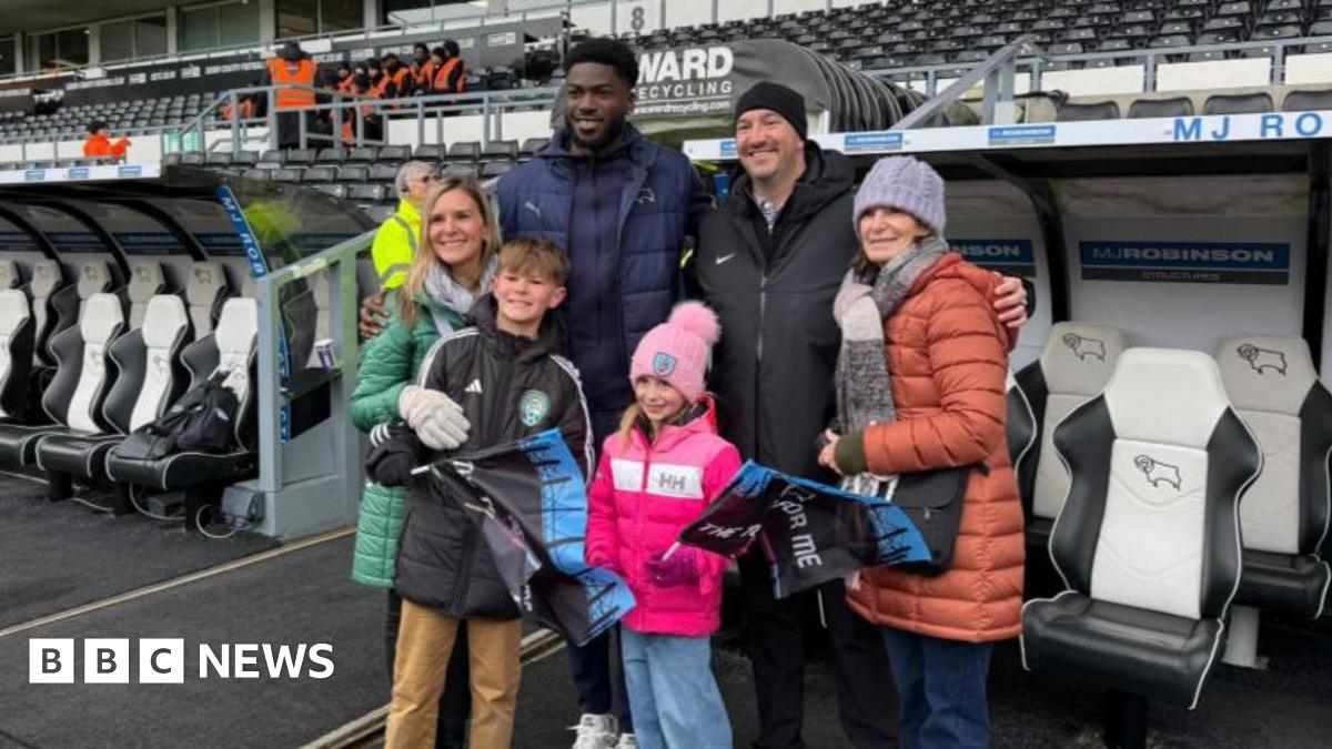Derby County player Patrick Agyemang flanked by two women, a man and two children. They are wearing thick coats and hats. They are stood in front of the dugouts pitch side.