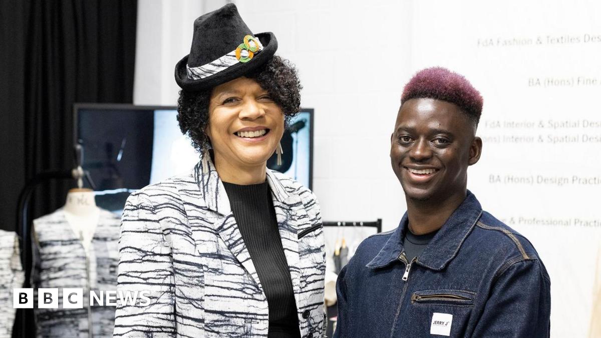 Chi Onwurah wearing a black and white, almost tiger-print suit jacket over a black top. She is wearing a small black hat with a strip of fabric and two orange and green circles. She is smiling into the camera and standing to the right of Lamin Gibba who has pink hair and is wearing a denim jacket. He is also smiling into the camera