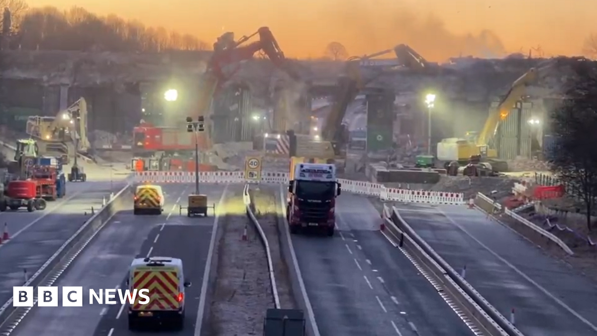 The eight lanes of the motorway are closed while multiple cranes work to pull down the concrete bridge overhead. Artificial work lights illuminate the dust rising from the works as the area is cordoned off behind plastic barriers. Vehicles are parked in front including two red and yellow highways' vans and a lorry.