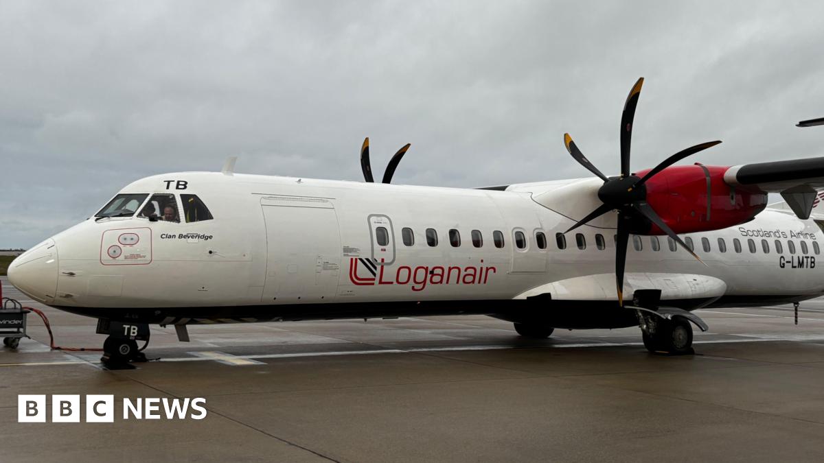 A shoulder shot photo of a man wearing a navy top wearing glasses standing in front of a pink and light brown government building.