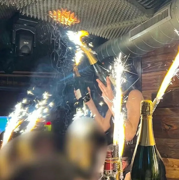 A waitress and partygoers holding sparkler-topped champagne bottles as the ceiling catches fire