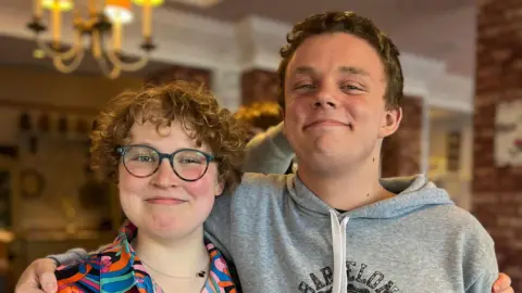 Emma Christie A young man stands with his arm around a teenage girl. They are both smiling broadly at the camera. Hazel is wearing glasses and a colourful shirt; her hair is short and curly. Lachlan has short hair and is wearing a grey hooded jumper. They appear to be in some kind of restaurant. 