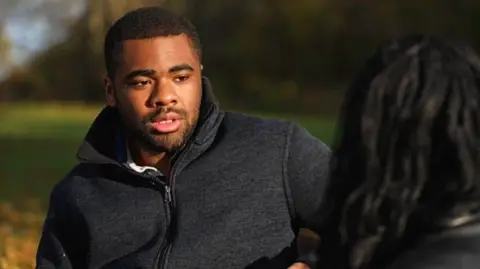 A young black man in a grey jacket sits on a park bench, speaking to a woman with long dark hair, whose face is obscured
