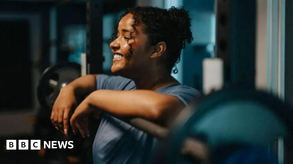 A woman with curly brown hair is in a gym, leaning on a weight. She is wearing a grey t-shirt and smiling