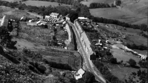 Gelligaer Historical Society Black and white image of a small village along the A469 road. A long row of terraced house can be seen from above stretching along either side of the road. Beyond that is predominantly farmland which incudes fields and trees on the side of a mountain.