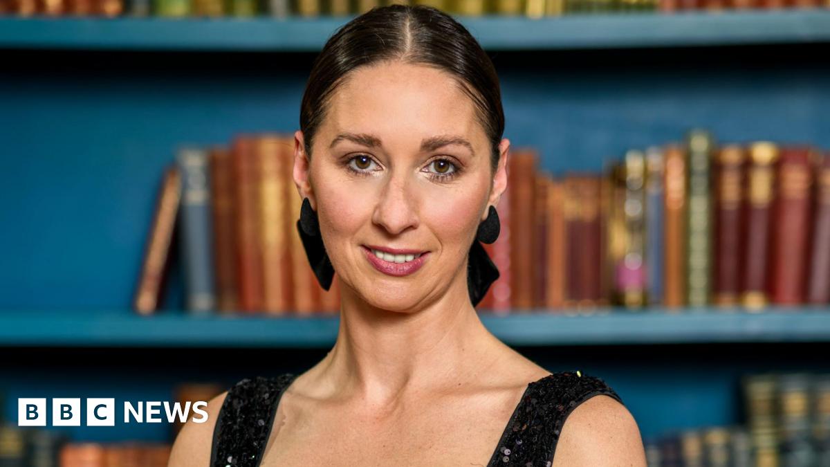 Rachel wearing a black sequined dress, her dark hair is tied up in a black bow. She is standing in front of a blue bookcase with different coloured books.