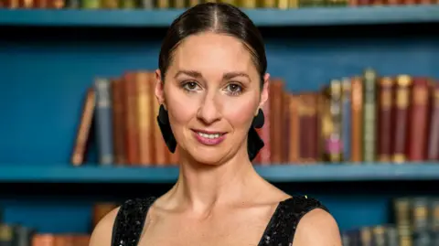 PA Media Rachel wearing a black sequined dress, her dark hair is tied up in a black bow. She is standing in front of a blue bookcase with different coloured books. 