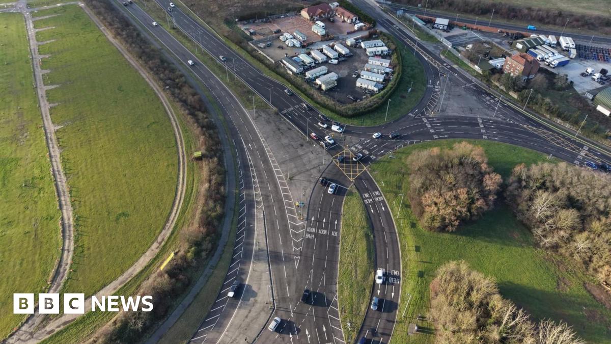 An aerial shot of a roundabout which is in the bottom right of the image and has trees planted on it. A dual carriageway is on the left of the image and merges on to the roundabout.
