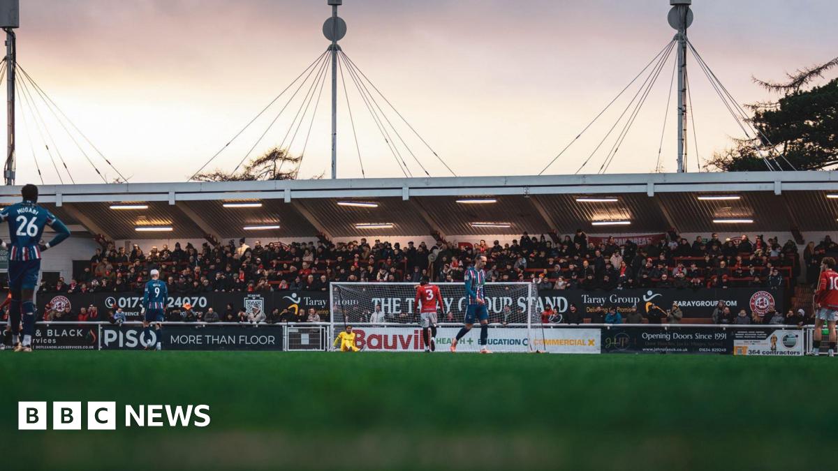Players from Chatham Town, in a red kit, and Carlisle United, in a blue kit, play on the pitch in front of a stand at the Bauvill Stadium.