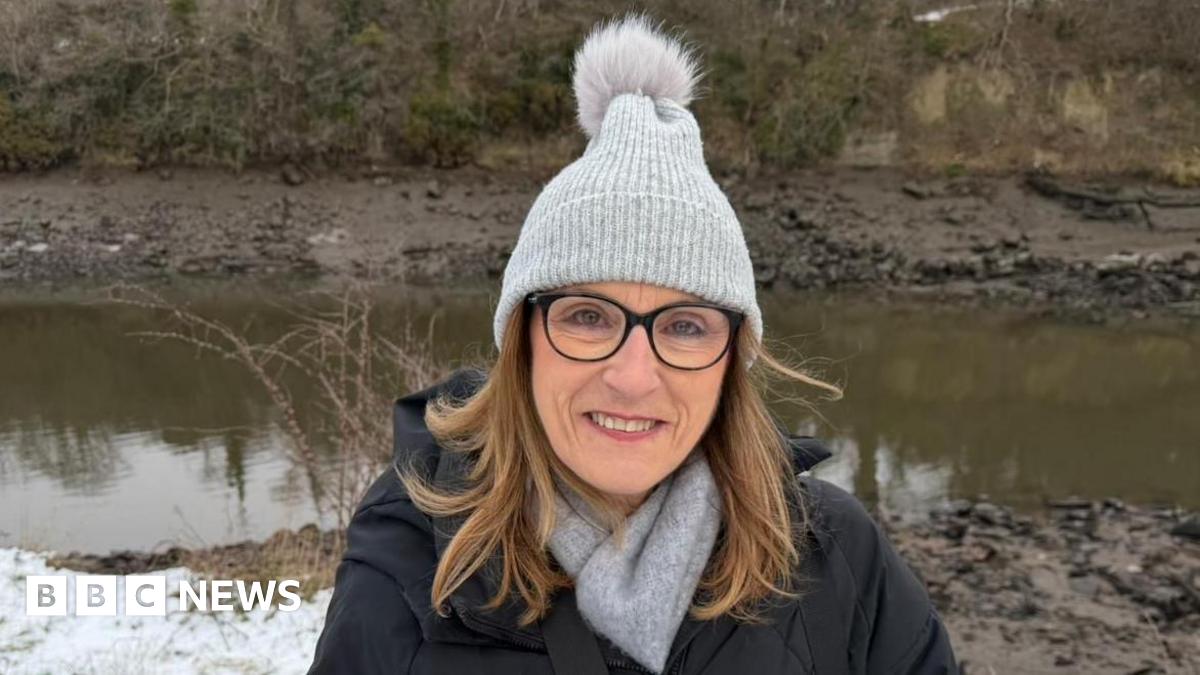 Allyson Tim standing by the edge of the River Wear. She has shoulder-length blonde hari and is wearing dark-rimmed glasses, a grey scarf and bobble hat. Snow can be seen behind her close to the river.