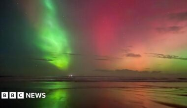 Green, pink, orange and pale yellow light glows above the beach and is reflected in the sea and wet sand.