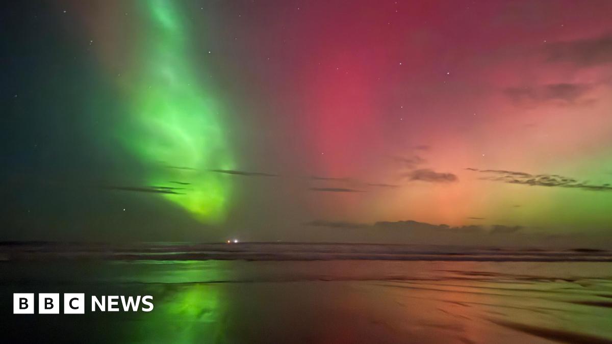 Green, pink, orange and pale yellow light glows above the beach and is reflected in the sea and wet sand.