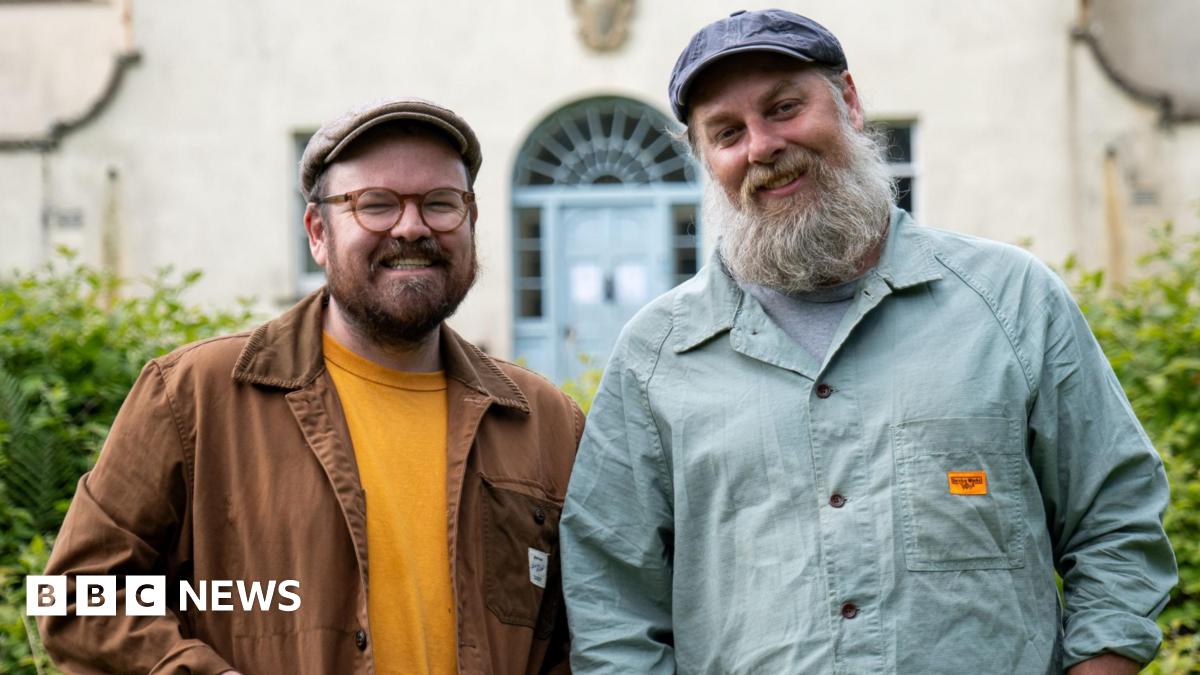 Banjo Beale and his husband Ro stand in front of the derelict mansion Ulva House, a grand but shabby building - its pale blue front door visible behind them. Banjo, with his trademark beard, glasses and flat cap, smiles alongside Ro,  also in a cap, who has a greying long beard.