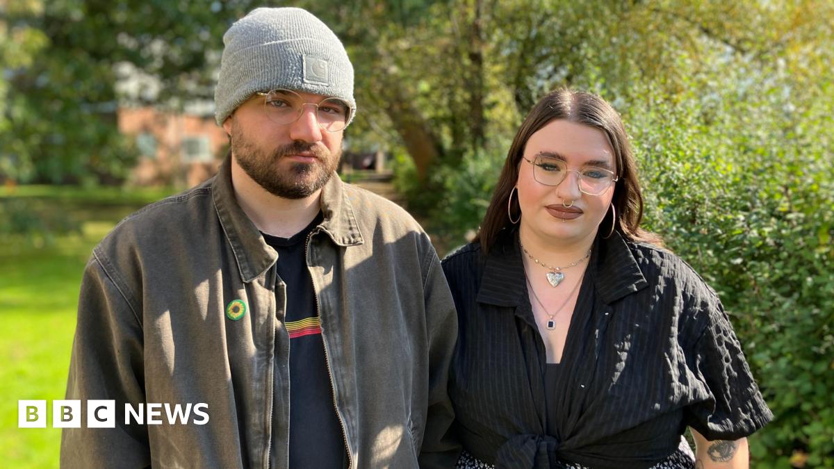 A man on the left with glasses, a grey beanie, and a short brown beard is wearing a light brown shirt open over a dark t-shirt. A woman next to him on the right also has glasses and is dressed all in black with long brown hair. They are standing outside with trees in the background.
