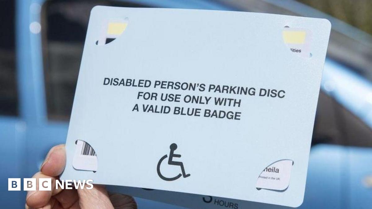 A hand holding a blue badge. On the front of the badge it says, DISABLED PERSON'S PARKING DISC FOR USE ONLY WITH A VALID BLUE BADGE. In the background of the picture is a blue car.