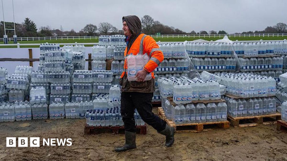 Millie Bobby Brown and David Harbour in character as Eleven and Jim Hopper. Eleven wears red shorts over grey leggings and a grey jumper. Jim Hopper wears a black leather jacket, a black beanie hat and dark trousers.They are speaking to director Frank Darabont on the set of Stranger Things. There is a vintage advertisement posted in the background with debrief on the ground.