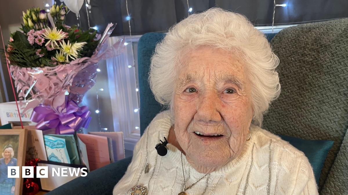Elsie Wilkins sits in a chair. She has short white hair and is wearing a white cable knit jumper. To her left there are flowers and cards.