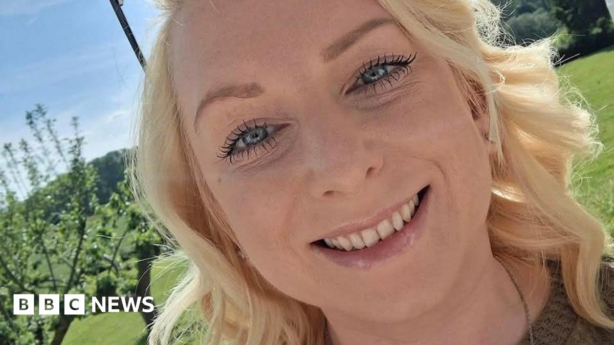 A blonde haired girl smiles at the camera. It is sunny and green in the background